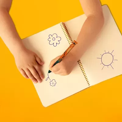 Caderno aberto com desenhos de flores e sol, mãos infantis a desenhar com lápis colorido sobre superfície amarela