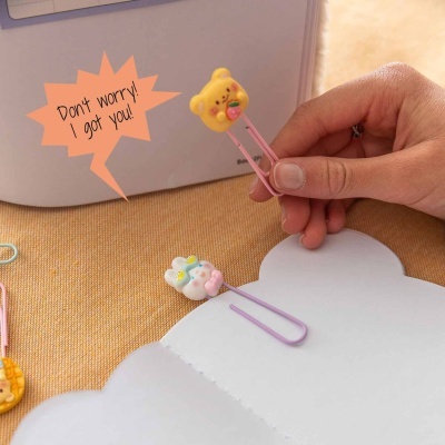Clipe de papel decorativo com figuras de ursinho e coelho, sobre tecido castanho claro e caderno branco.