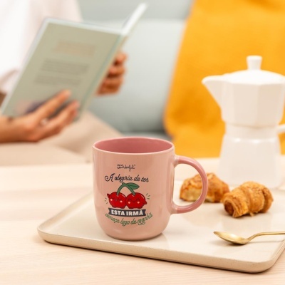 Caneca rosa com desenho de cerejas e texto na bandeja com croissants, colher e cafeteira