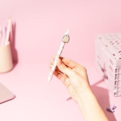 Caneta branca com desenhos de gato e capuz de borracha em forma de gato segurada à frente de fundo rosa