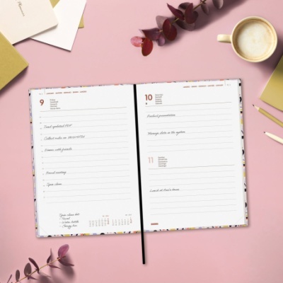 Agenda aberta sobre mesa rosa com texto manuscrito, chávena de café, folhas secas e lápis