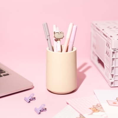 Porta-canetas bege com canetas coloridas e marcador de gato sobre mesa rosa com laptop e clips lilás