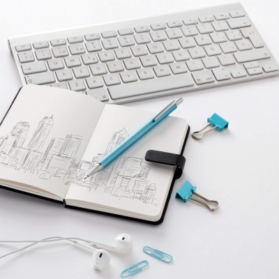 Mesa com teclado branco, caderno com desenho de edifícios, caneta azul, clips e auscultadores brancos