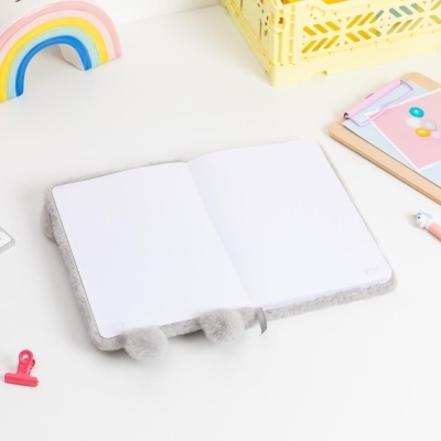 Caderno cinzento felpudo aberto com pompons, caneta unicórnio, bloco de notas colorido, clipe vermelho e arco-íris decorativo em madeira.