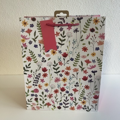 Saco de presente em papel com padrão colorido de flores e folhas e etiqueta rosa.
