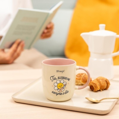 Caneca bege com interior e asa rosa, texto e flor desenhada, em tabuleiro com croissants e cafeteira branca