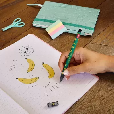 Mão a escrever com caneta verde decorada com bananas num caderno riscado com desenho de macaco e bananas, estojo azul claro, post-its coloridos e tesoura azul clara numa mesa de madeira.
