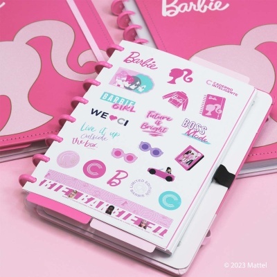 Caderno branco com desenhos e texto coloridos da Barbie e acabamento em anéis rosa numa mesa rosa