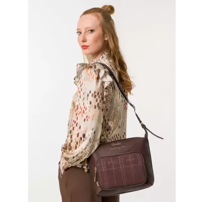 Mulher com mala de ombro castanha, bolso xadrez frontal, alça preta, camisa colorida e calças castanhas