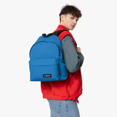 Mochila azul da marca EASTPAK usada por pessoa com colete vermelho e camisa cinza.