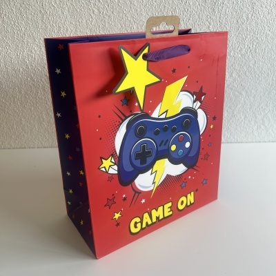 Saco de presente vermelho com ilustração de comando de videojogo azul e texto GAME ON