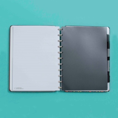 Caderno aberto com folhas brancas pautadas e capa cinza escuro sobre fundo azul.