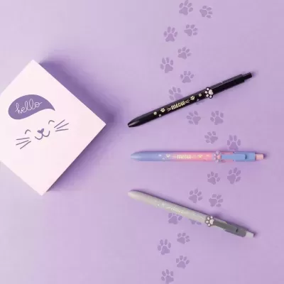 Bloco de notas com rosto de gato e três canetas coloridas com tema de gato sobre superfície lilás com pegadas de gato.