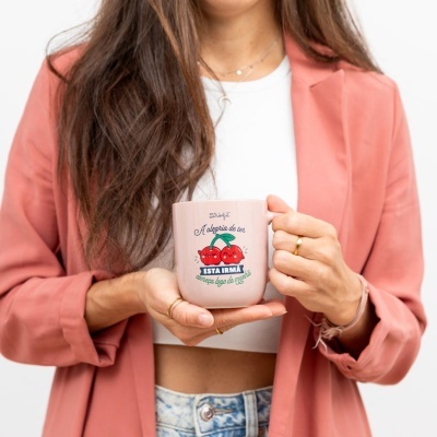Caneca rosa com design de cerejas e texto em português, segurada por mulher vestindo casaco rosa e top branco.