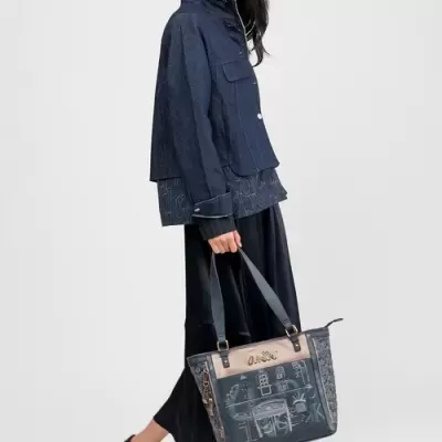 Mulher com casaco azul escuro, calças pretas largas, sapatos pretos e mala azul com desenhos e texto Anekke