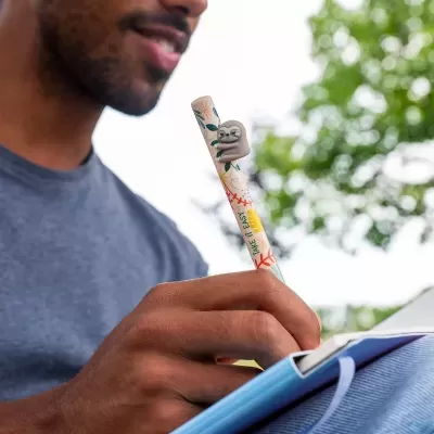 Caneta decorada com figura de preguiça e texto TAKE IT EASY LOVE, homem a escrever num caderno azul ao ar livre