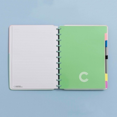Caderno aberto com capa verde, páginas pautadas e separadores coloridos sobre fundo azul claro