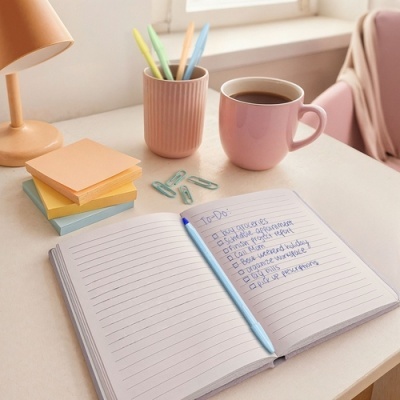 Mesa com bloco de notas aberto, caneta azul, caneca cor-de-rosa, suporte com canetas coloridas, post-its e clips