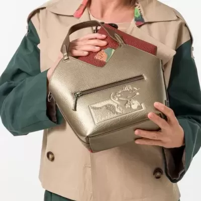 Bolsa de mão dourada com bolso frontal zip e personagem em relevo, segurada por pessoa com casaco bege e verde.