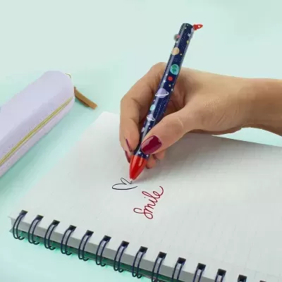 Mão a escrever 'Smile' com caneta decorada com tema espacial num caderno de anel espiral