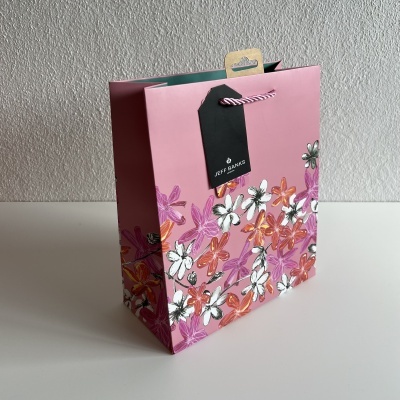Saco de presente rosa com flores brancas, laranja e lilás e etiqueta preta com cordão.