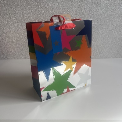 Saco de papel colorido com estrelas e alças vermelhas numa superfície branca