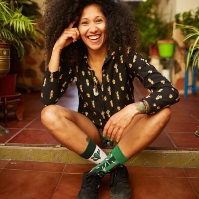 Pessoa com camisa preta estampada de cactos, meias verdes com cactos, sapatos pretos, sentada em chão de azulejo vermelho com plantas ao fundo