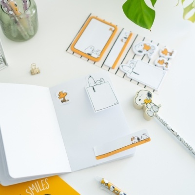 Notas autocolantes e marcadores de página com Snoopy e Woodstock, mesa branca com canetas e folhas.