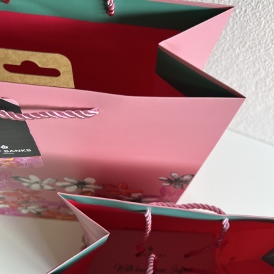 Sacos de papel rosa e vermelho com alças torcidas rosa, sobre superfície branca com parede ao fundo.