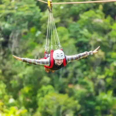 Pessoa a usar capacete e colete suspensa em tirolesa com vegetação ao fundo