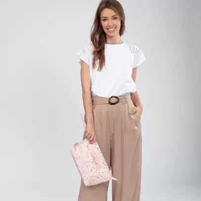 Mulher com blusa branca rendada e calças camel segurando bolsa rosa clara com padrão negro