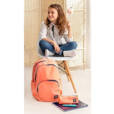 Mochila cor de salmão com estojo e caderno em primeiro plano e menina sentada numa cadeira atrás