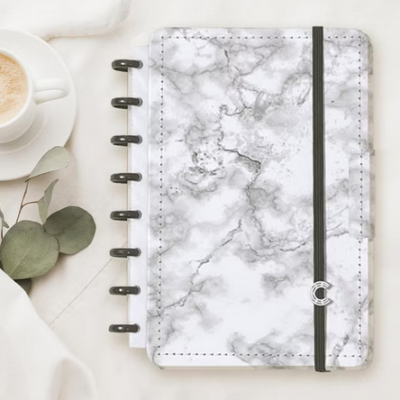 Caderno com capa de mármore cinzento e branco com elástico preto, junto a café e folhas verdes