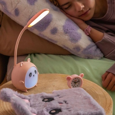 Lâmpada de mesa em forma de panda cor-de-rosa com luz ligada, despertador cor-de-rosa BZ-F, almofada manchada e capa cinza peluda numa mesa redonda de madeira
