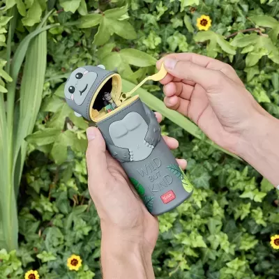 Estojo em forma de gorila cinzento com zíper amarelo aberto, com folhagem e flores ao fundo.