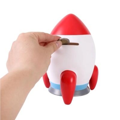 Mealheiro em forma de foguetão com partes vermelhas e brancas, com mão a inserir moeda
