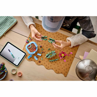 Pessoa a montar brinquedo de construção de peças coloridas sobre tapete castanho com tablet com instruções numa mesa de madeira clara