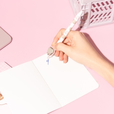 Caneta decorativa branca com gatinhos a escrever em papel branco sobre mesa rosa