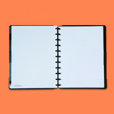 Caderno preto aberto com páginas internas brancas, uma riscada, outra em branco, sobre fundo laranja.