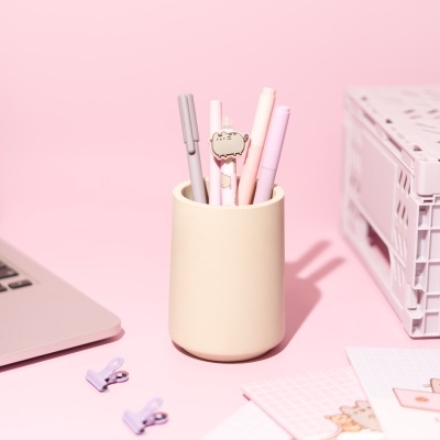 Canetas em organizador de mesa bege com detalhes em rosa e lilás, mesa rosa com portátil e acessórios de escritório.
