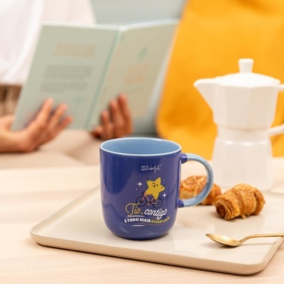 Caneca azul com texto e desenho de estrela a andar de bicicleta numa bandeja com croissants e colher dourada