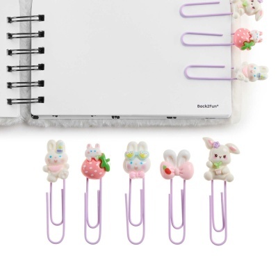 Clips de papel cor-de-rosa com figuras de coelhos e morangos, ao lado de um caderno branco com capa peluda.