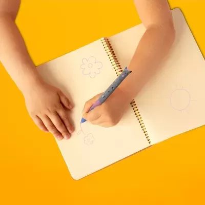 Criança desenhando flores num caderno espiral com caneta em forma de baleia sobre fundo amarelo