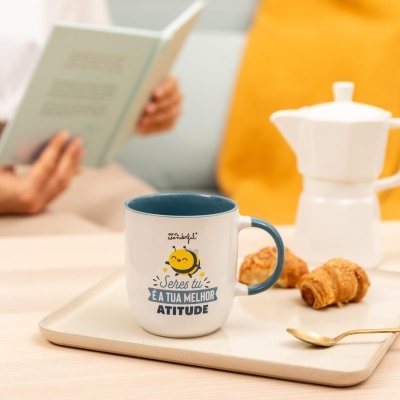 Caneca com desenho de abelha e texto inspirador, croissants num tabuleiro e uma cafeteira ao fundo