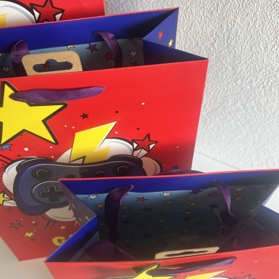 Sacolas de papel vermelha e azul com alças de fita roxa e estampas de estrelas e controle de videojogo