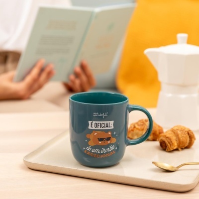 Caneca azul com texto e desenho de urso, croissants, prato e pessoa a ler livro.