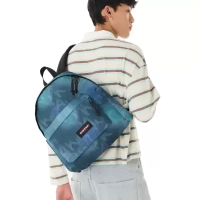 Mochila azul EASTPAK com padrão, bolso frontal e alças pretas numa pessoa com camisa às riscas e calças cinza.