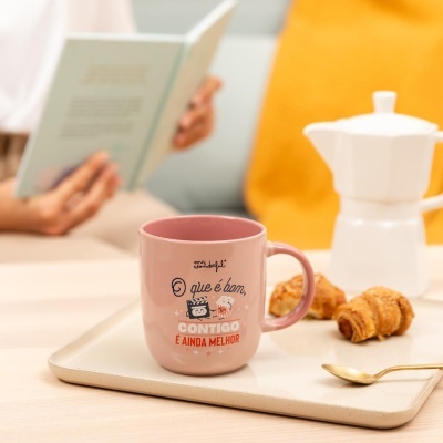 Caneca rosa com frase inspiradora sobre uma bandeja com croissants e cafeteira, pessoa lendo livro ao fundo.