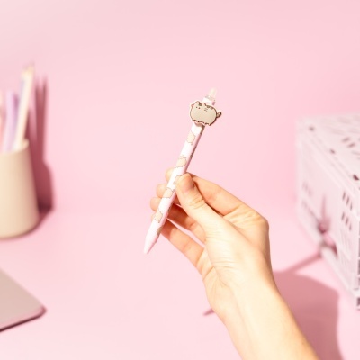 Caneta rosa com pequenos gatos e adorno de gato no topo, segurada por uma mão