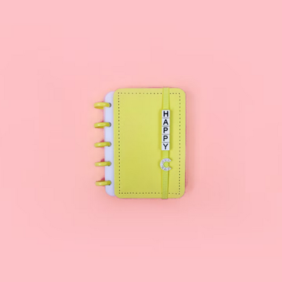Caderno amarelo com argolas e palavra HAPPY em blocos brancos sobre fundo cor-de-rosa.